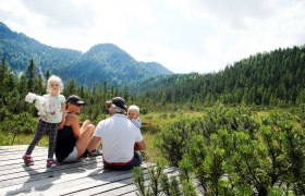 High moor Leckermoos, © weinfranz.at Family sitting on a wooden platform in the Leckermoos high moor, surrounded by mountains and forest.