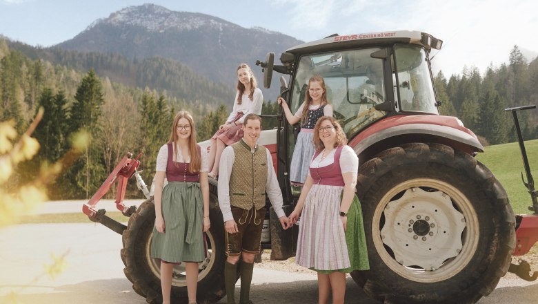Gastgeberfamilie, © grasberger Eine Familie in traditioneller Tracht steht vor einem Traktor in einer ländlichen Berglandschaft.