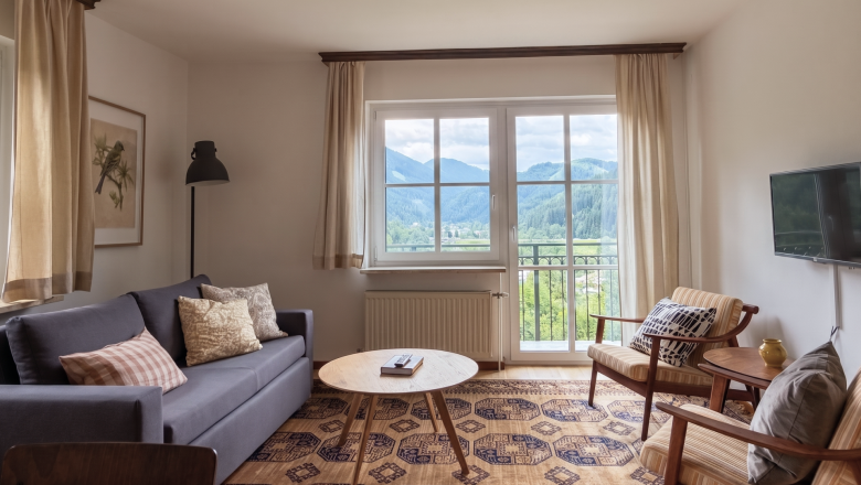 Lina Wohnen Hero, © Raphaela Bachler Cozy living room with a sofa, armchairs, and a large window offering a view of the mountains.