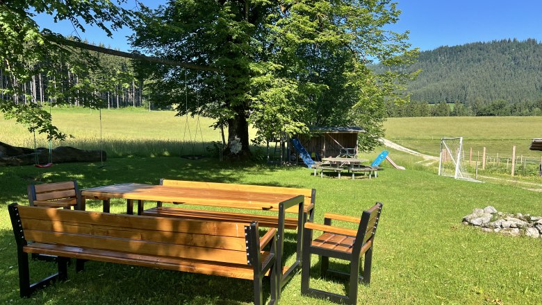 Garten der Ferienwohnung am Biohof Herdengl, © Ybbstaler Alpen_Karin Hager Ein Garten mit Holztisch und Bänken, umgeben von Bäumen und Wiesen, mit einem Spielplatz im Hintergrund.