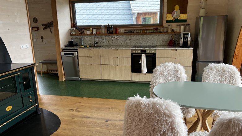 Biohof Oberhof (c)Karin Hager Ybbstaler Alpen (1), © Ybbstaler Alpen_Karin Hager Modern kitchen with wood paneling, green stove, dining table with fluffy chairs and large window with view to the outside.