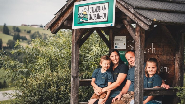 Fotoschooting_Panoramahöhenweg-jpg_3, © Familie Korntheuer- Fotoshooting Panoramahöhenweg Familie Korntheuer sitzt in einem Holzunterstand mit einem Schild 'Urlaub am Bauernhof'. Im Hintergrund sind grüne Hügel zu sehen.