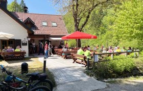 Schindlhütte im Sommer, © Andreas Rasch Eine Berghütte mit Terrasse, auf der Menschen an Tischen sitzen. Fahrräder sind im Vordergrund zu sehen, umgeben von grüner Natur.