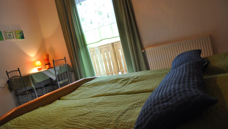 Fuchsbauernhof, © Fuchsbauernhof Gemütliches Schlafzimmer mit grünem Bett, Tisch und Stühlen, Lampe und Fenster mit Vorhängen.