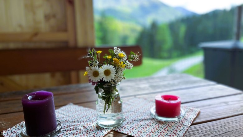 Gastgarten genießen, © grasberger Ein Holztisch mit zwei Kerzen und einem kleinen Blumenstrauß in einem Glasgefäß, im Hintergrund eine verschwommene Berglandschaft.