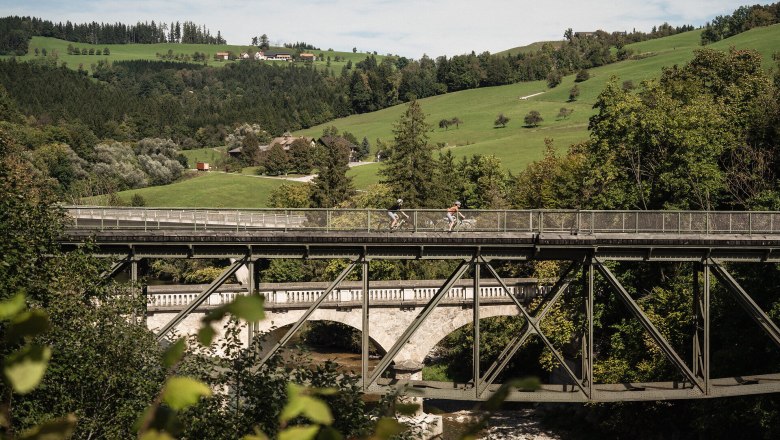 Ybbstalradweg, Rad fahren, Gravel, Mostviertel, © Niederösterreich Werbung/www.tomasslavik.cz Das Bild zeigt eine idyllische Landschaft entlang des Ybbstalradwegs, in der zwei Radfahrer:innen über eine Brücke fahren, umgeben von grünen Wiesen, Bäumen und einem klaren Himmel, der ein Gefühl von Freiheit und Naturverbundenheit vermittelt.