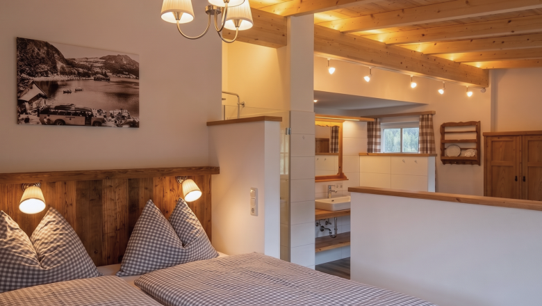 Amon Schlafzimmer und Bad, © Raphaela Bachler A cozy bedroom with a wooden beam ceiling and a double bed, decorated with checkered pillows and blankets. In the background is a bathroom with a sink; behind the half-height wall is a freestanding bathtub.