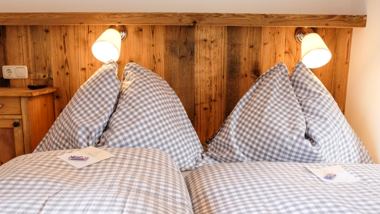 Amon Schlafzimmer, © Raphaela Bachler A cozy double bed with checkered duvet covers and two reading lamps mounted on a wooden headboard.