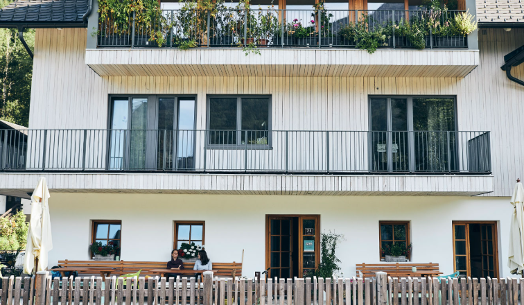 Das Haus am See in Lunz, © Familie Mayr Ein modernes dreistöckiges Haus mit Holzbalkonen und Pflanzen. Vor dem Haus sitzen zwei Personen auf einer Bank hinter einem Holzzaun.