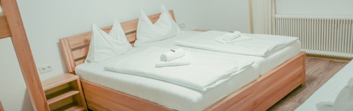 JoSchi Almgasthof Hochkar, © Martin Stellenberger/JoSchi Hochkar GmbH Ein gemütliches Doppelbett in einem hellen Zimmer mit Holzrahmen und weißen Bettlaken, dekoriert mit gefalteten Handtüchern.