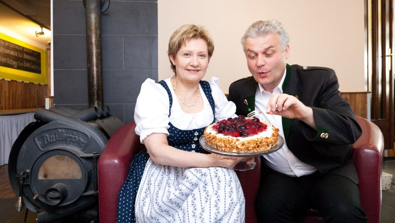 Gasthof Ettel, © Gerald Lechner Ein Mann und eine Frau in traditioneller Kleidung halten eine Torte vor einem Ofen.