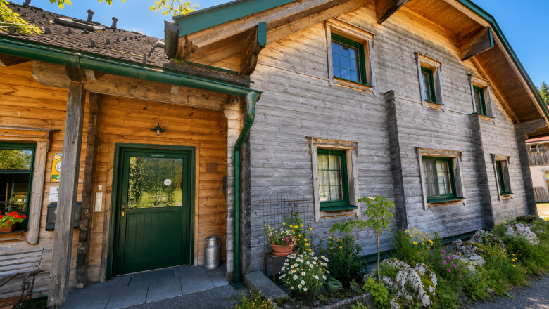 Biohof Herdengl, © Ybbstaler Alpen_Karin Hager Ein traditionelles Holzhaus mit grünen Fensterrahmen und einer grünen Tür, umgeben von Blumen und Pflanzen, unter einem klaren blauen Himmel.