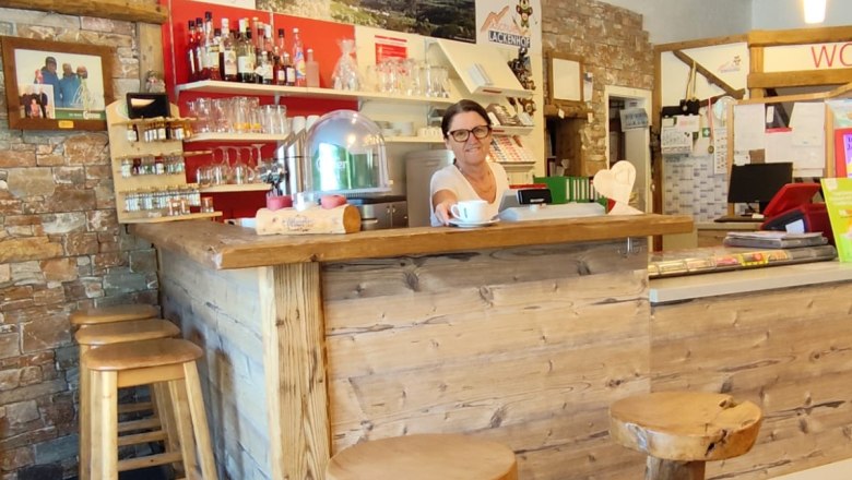 edelweisslounge-cfranz-heher, © Franz Heher A woman stands behind a rustic wooden counter in a cozy lounge with wooden stools and a stone wall.