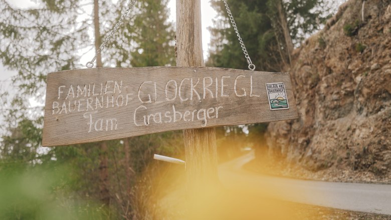 Ankommen am Bauernhof, © Auftragsfoto.at Holzschild mit der Aufschrift 'Familienbauernhof Glockriegl Fam. Grasberger' an einem Waldweg.