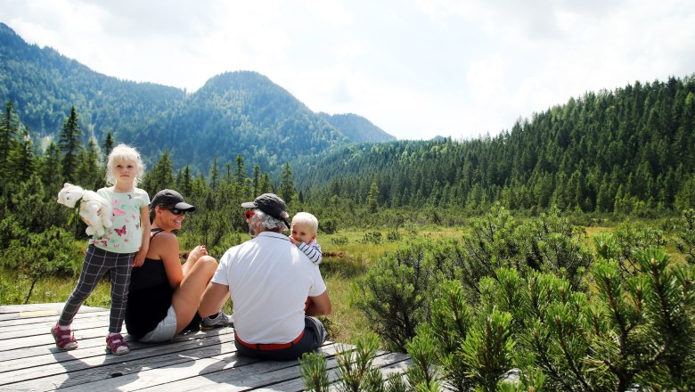 High moor Leckermoos, © weinfranz.at Family sitting on a wooden platform in the Leckermoos high moor, surrounded by mountains and forest.