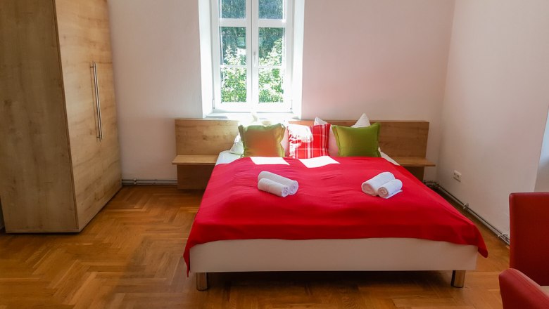 kuscheliges Schlafzimmer, © Monika Stock Schlafzimmer mit rotem Bettüberwurf, Kissen und Handtüchern auf dem Bett, Holzschrank und Fenster im Hintergrund.