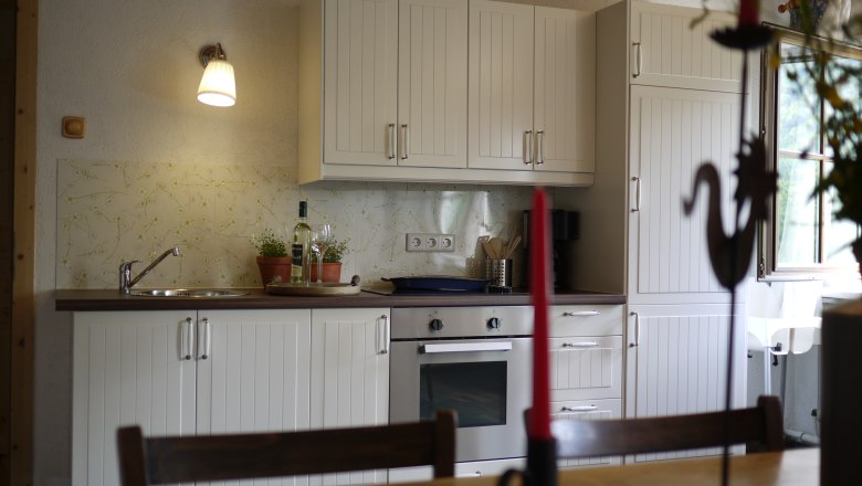 Rinntaverne, © Rinntaverne Küche mit weißen Schränken, Herd und Spüle, dekoriert mit Pflanzen und einer Weinflasche.