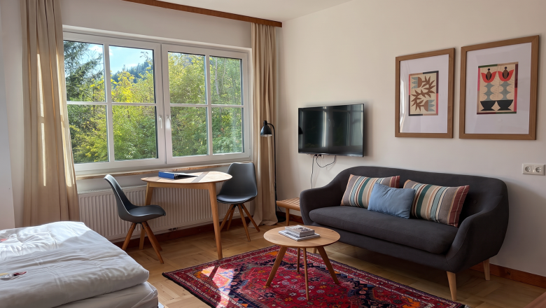 Magda Couch, © Raphaela Bachler Cozy living room with a sofa, table, chairs, and a large window overlooking the greenery.