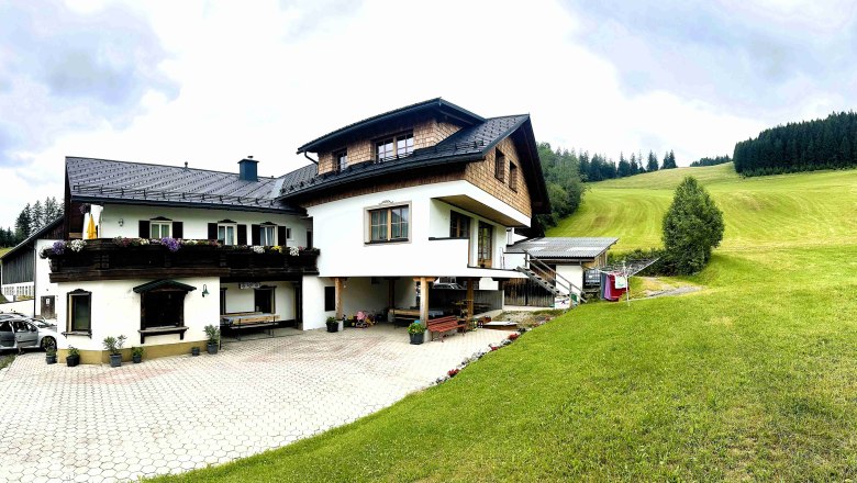 image00023, © Familie Buber A traditional farmhouse with wood siding and flowers on the balcony, surrounded by green meadows and forests.