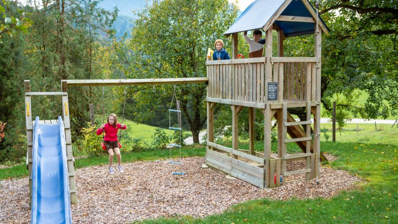 Spielplatz, © grasberger Kinder spielen auf einem Spielplatz mit Schaukel, Rutsche und Kletterturm in einer grünen Umgebung.
