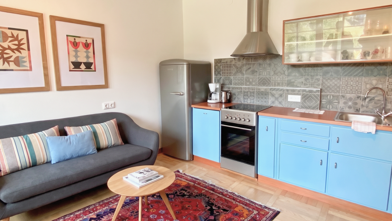 Magda Overview, © Raphaela Bachler A cozy kitchen with blue cabinets, a gray sofa, and a colorful rug.