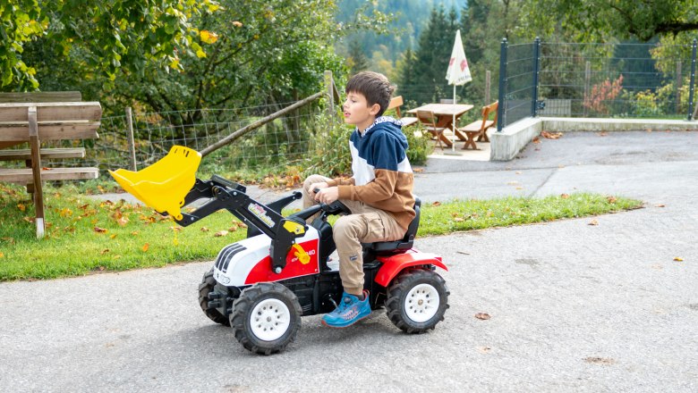 Trettraktor für die Kids, © grasberger Ein Kind fährt auf einem kleinen Trettraktor mit Schaufel auf einem Weg im Freien.