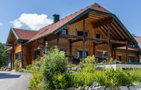 Biohof Herdengl in Lunz am See, © Theo Kust Ein traditionelles Holzhaus mit einem roten Dach, umgeben von bunten Blumen und grünen Pflanzen, unter einem blauen Himmel.