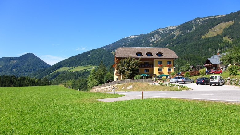 Schneeschuh Wandern, © Gasthof Stiegenwirt Ein gelbes Gebäude mit Balkon und Blumen, umgeben von grünen Wiesen und Bergen unter blauem Himmel.