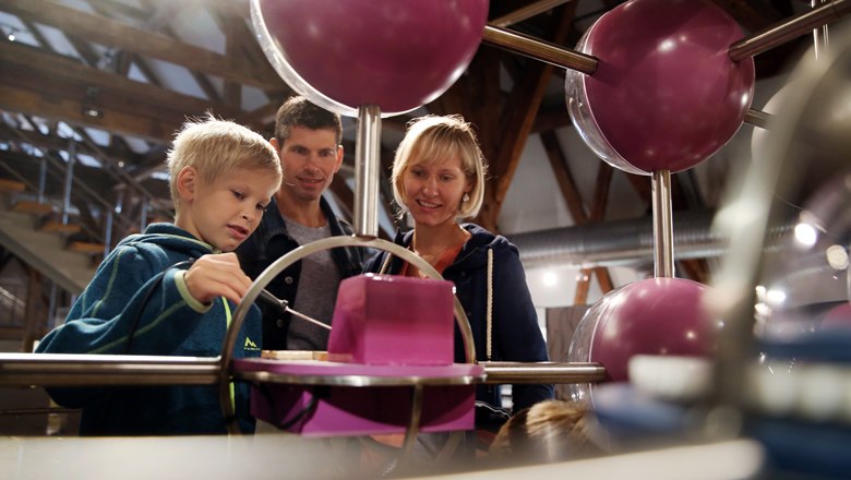 FeRRum - Welt des Eisens, © weinfranz.at Eine Familie betrachtet eine interaktive Ausstellung mit großen, violetten Kugeln.