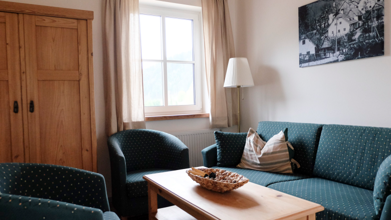 Amon Couch, © Raphaela Bachler A cozy living room with green upholstered furniture, a wooden table, and a window with curtains.