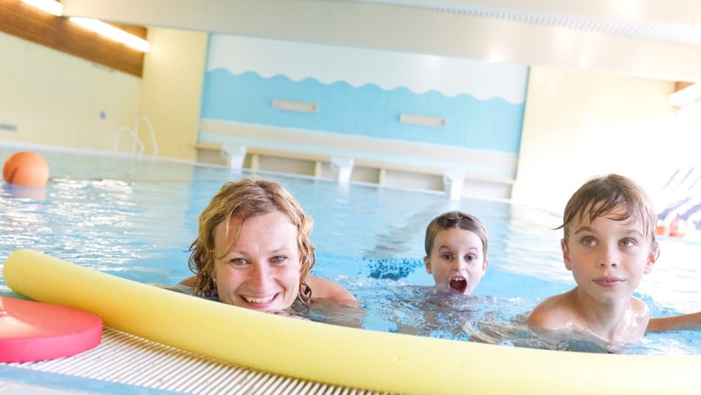 Bathing family, © Theo Kust A woman and two children swim in an indoor pool, smiling and playing with a yellow pool noodle.