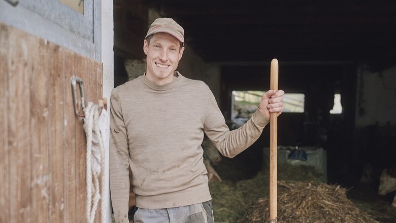 Bauer Peter bei der Arbeit, © Auftragsfoto.at Ein lächelnder Mann in Arbeitskleidung steht in einem Stall und hält eine Heugabel.