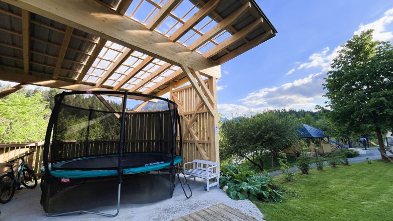 Überdachtes Trampolin, © grasberger Überdachtes Trampolin in einem Garten mit Holzdach und umgebender Natur.