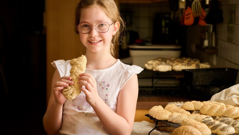 Frisch schmeckts am Besten, © grasberger Ein Mädchen mit Brille hält ein Gebäckstück in der Hand und lächelt in die Kamera. Im Hintergrund sind weitere Backwaren auf einem Tisch zu sehen.