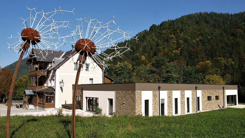 Fastenhaus Maria Seesal, © Fasthaus Maria Seesal Ein modernes Gebäude mit großen Löwenzahn-Skulpturen im Vordergrund, umgeben von grünen Hügeln und Bäumen.