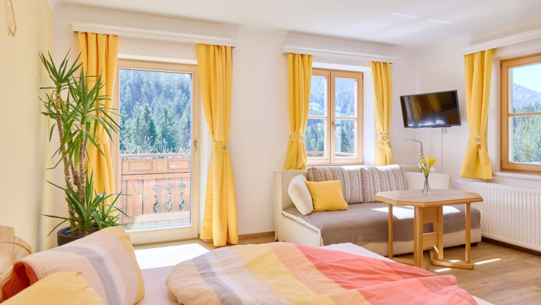 Sonnige Ferienwohnung 1 - Schlaf- und Wohnbereich, © grasberger Heller Raum mit Bett, Sofa, Tisch und gelben Vorhängen, Blick auf Wald und Berge.
