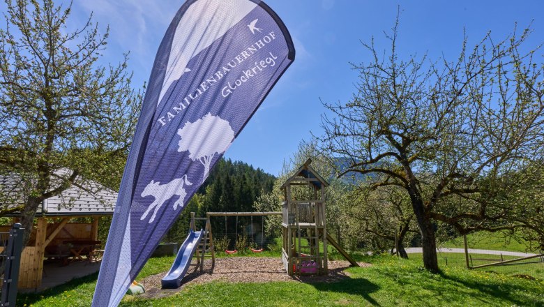 Gästegarten mit herrlicher Aussicht, © grasberger Spielplatz mit Rutsche und Schaukel im Garten eines Bauernhofs, umgeben von Bäumen und Wiesen.