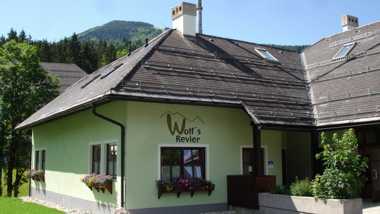 Wolf´s Revier, © Robert Wolf Ein grünes Gebäude mit der Aufschrift 'Wolf's Revier' und Blumenkästen vor den Fenstern, umgeben von Bäumen und Bergen im Hintergrund.