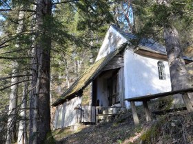 Eine kleine wei&szlig;e Kapelle im Wald, umgeben von B&auml;umen und einem Holztisch im Vordergrund.