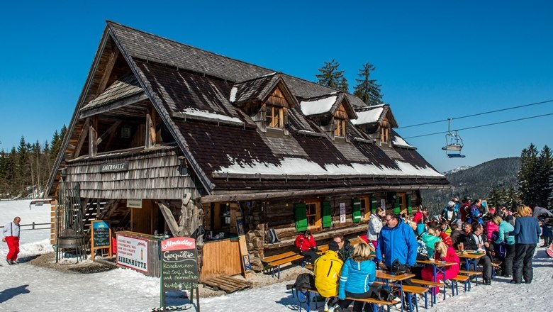 A mountain hut in the snow with people sitting outside.