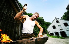 Ein Schmied arbeitet mit einem Hammer auf einem Amboss vor einem Gebäude im Freien.