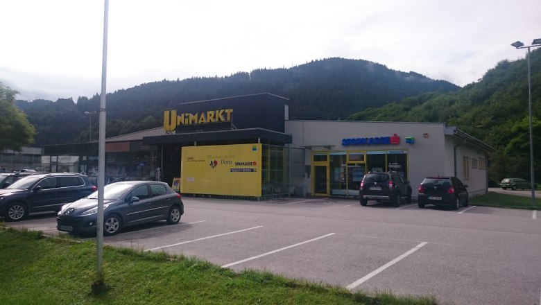 Shopping center with Unimarkt and Sparkasse in front of wooded hills.