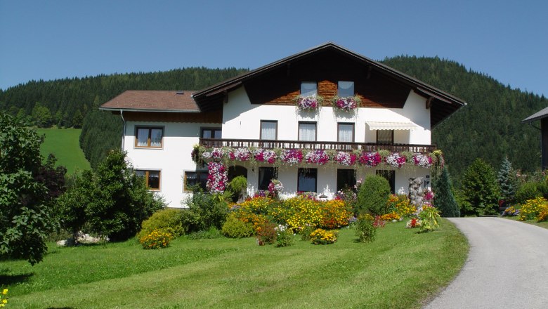 Ein weißes Haus mit Blumenbalkonen vor einem bewaldeten Hügel.