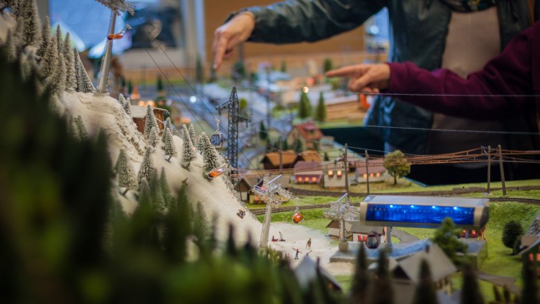 Detailreiche Modelllandschaft mit Seilbahn und schneebedeckten Bäumen, im Hintergrund zeigen zwei Personen auf das Modell.
