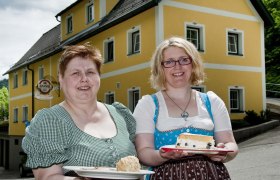 Zwei Frauen in traditioneller Kleidung halten Teller mit Speisen vor einem gelben Gebäude.