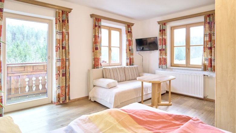 Helles Zimmer mit Sofa, Tisch, Fernseher und Balkon mit Waldblick.