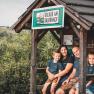 Familie Korntheuer sitzt in einem Holzunterstand mit einem Schild 'Urlaub am Bauernhof'. Im Hintergrund sind gr&uuml;ne H&uuml;gel zu sehen.