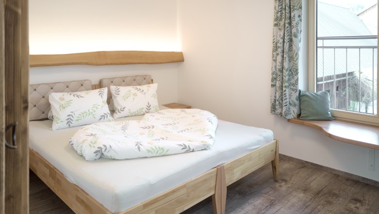 Ein gem&uuml;tliches Schlafzimmer mit einem Holzbett, dekoriert mit Pflanzenmotiven auf Bettw&auml;sche und Vorh&auml;ngen. Ein Fenster mit Sitzbank bietet Ausblick nach drau&szlig;en.