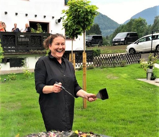 Eine Frau steht lachend an einer Feuerstelle im Freien, umgeben von gr&uuml;ner Landschaft und einem Geb&auml;ude im Hintergrund.