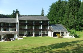 Hotel building with solar panels and green meadow in the foreground, surrounded by forest.